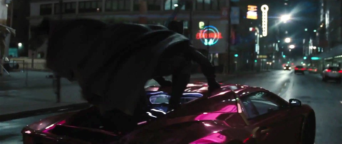 Batman di schiena accovacciato sul tettuccio di un'auto colorata