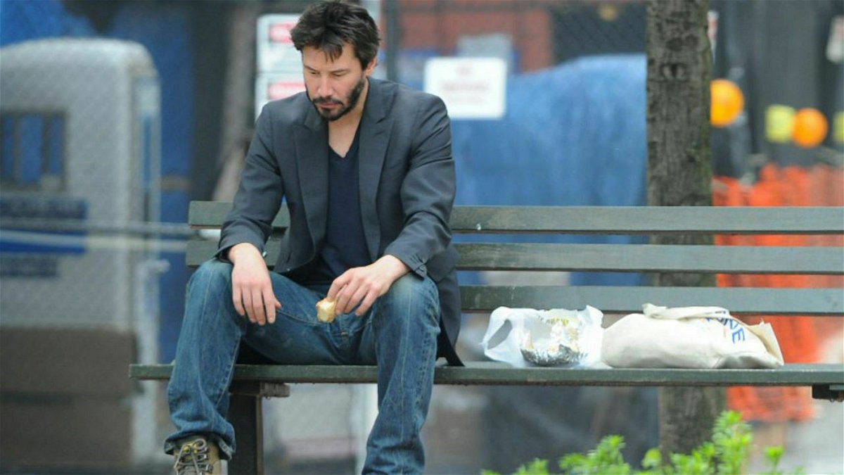L'attore Keanu Reeves mangia un panino seduto con aria abbattuta su una panchina